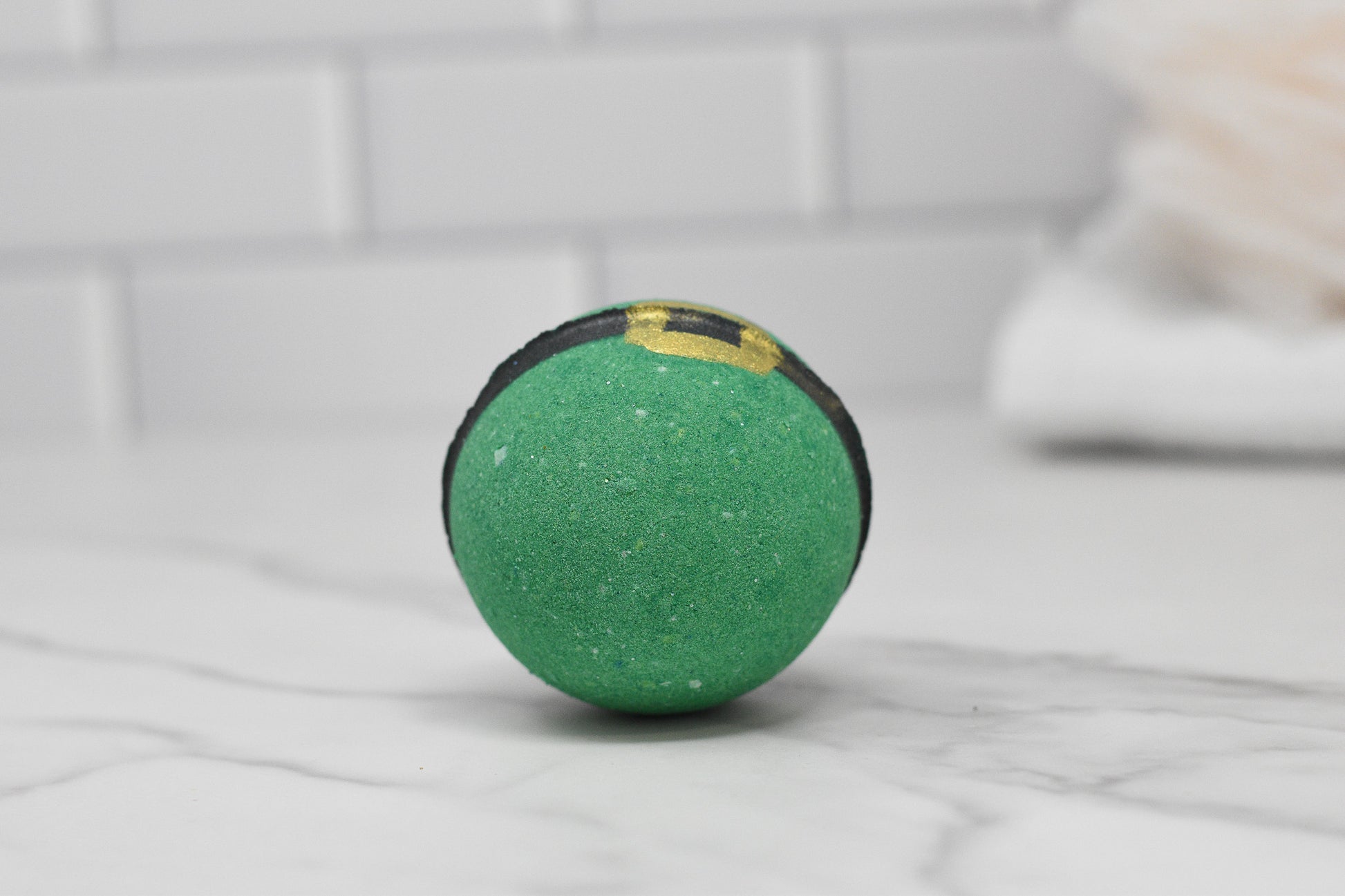 The Enchanting Elf Bath Bomb: Round by 570 Soap Co. presents a whimsical charm with its green color, black stripe, and gold buckle detailing as it rests on a marble surface. The scene is complemented by white subway tiles and a blurred towel in the background, making it perfect for skin-nourishing relaxation.