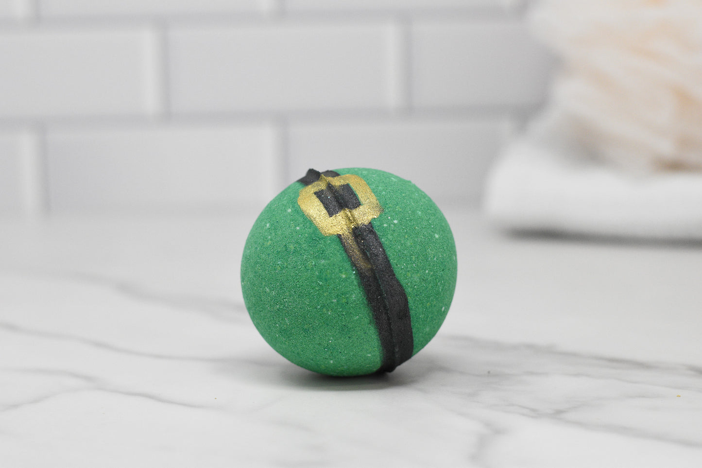 An Enchanting Elf Bath Bomb from 570 Soap Co., featuring a round shape with a black belt design and gold buckle, rests on a white marble surface, emanating whimsical charm. In the background, the allure of this skin-nourishing treat is enhanced by the soft blur of white subway tiles and a towel.