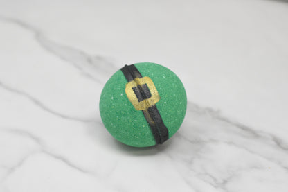 An Enchanting Elf Bath Bomb: Round, crafted by 570 Soap Co. and featuring a whimsical black and gold belt design reminiscent of a leprechaun's, rests on a marble surface, promising skin-nourishing magic.