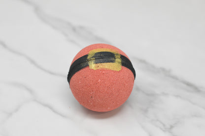 The Jolly Santa Bath Bomb by 570 Soap Co., a round, red creation resembling Santa's belt with its black band and gold buckle, releases an invigorating Orange Soda Pop fragrance while resting on a white marble surface.