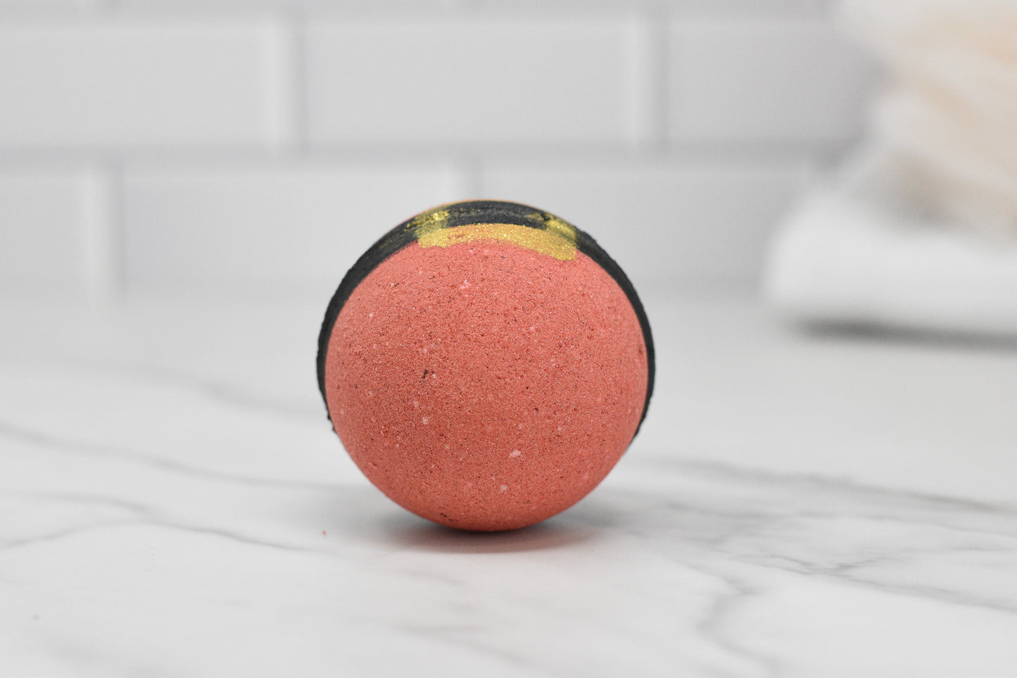 A Jolly Santa Bath Bomb: Round by 570 Soap Co., showcasing a vibrant orange color with black and yellow stripes, gracefully sits on a marble surface. With the refreshing aroma of Orange Soda Pop, it adds holiday cheer against the backdrop of a white tiled wall.