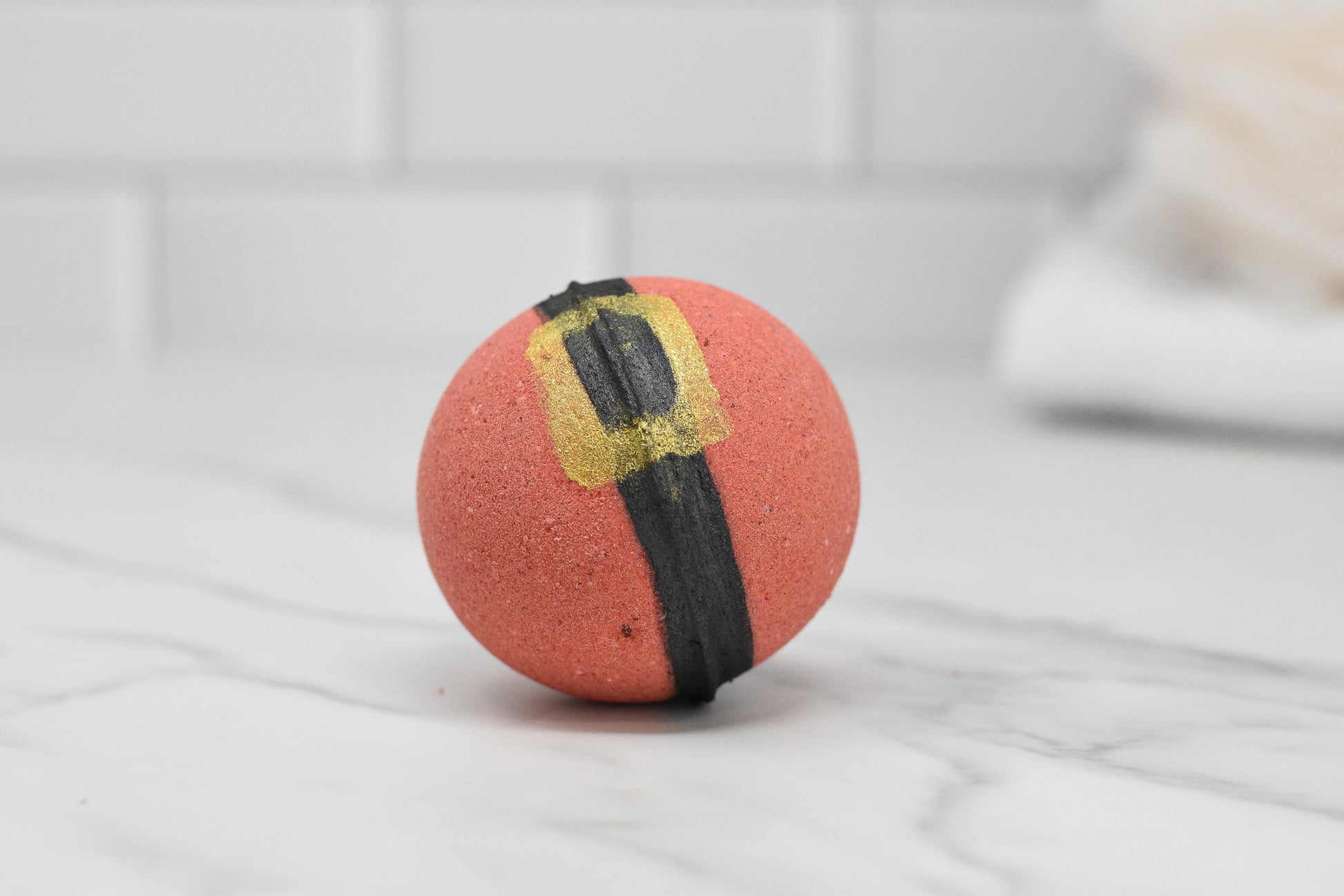 A red, round Jolly Santa Bath Bomb from 570 Soap Co. is displayed on a white marble surface, showcasing its black and gold belt design. The backdrop includes white, rectangular tiles and a blurred white towel, perfectly capturing the festive essence of a holiday bath bomb with its playful Orange Soda Pop fragrance.