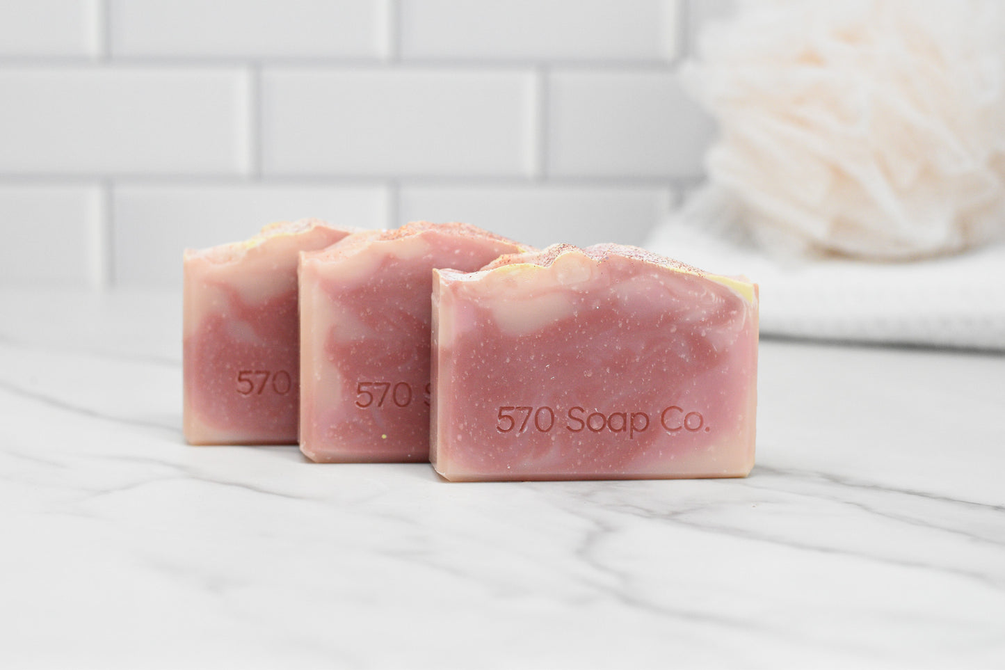 Three Sweet Serenade Soap Bars from 570 Soap Co., featuring an exotic fragrance, are elegantly placed on a white marble surface. The softly focused background accentuates the white tiled walls and a loofah, emphasizing the charm of natural ingredients in each pink marbled bar.