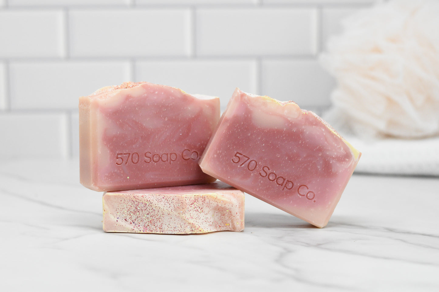 Three Sweet Serenade Soap Bars in pink and white from 570 Soap Co. are elegantly arranged on a white marble surface against a tiled backsplash, with a blurred bath sponge in the background suggesting the exotic fragrance and natural ingredients of each handcrafted bar.