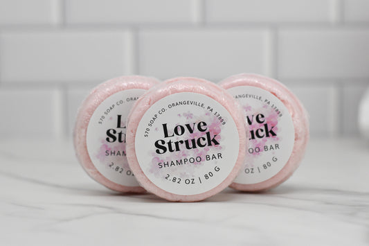 Three Love Struck Solid Shampoo Bars from 570 Soap Co., each weighing 2.82 oz / 80 g, are stacked on a marble countertop. Their pink circular design highlights your eco-friendly choice while chic white subway tiles in the background make them perfect for anyone curating a sustainable beauty routine.