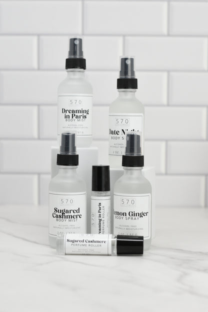 Displayed against a white tiled background, the collection from 570 Soap Co. includes their Luxurious Fragranced Body Mist Spray in scents like Dreaming in Paris, Sugared Cashmere, Date Night, and Lemon Ginger. These alcohol-free body sprays and perfume rollers feature hydrating technology for a refreshing experience.