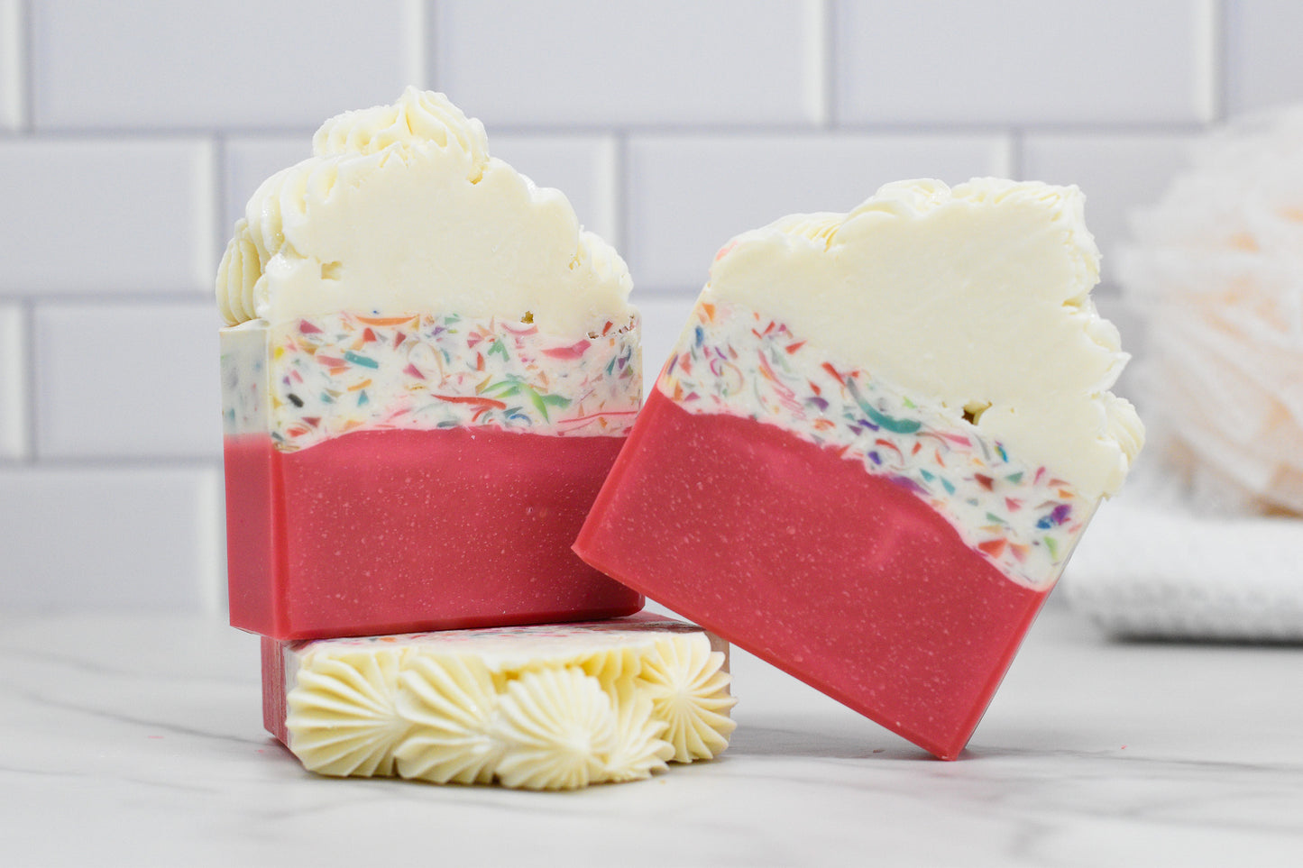 A pair of bars from the Birthday Cake Bar Soap collection by 570 Soap Co. exudes a buttercream icing fragrance, showcasing a white top layer adorned with confetti-like colors and a pink bottom. The soap is intricately textured on top, resembling birthday cake. One bar is leaned against the other, with a loofah softly blurred in the background.