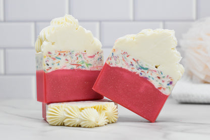 A pair of bars from the Birthday Cake Bar Soap collection by 570 Soap Co. exudes a buttercream icing fragrance, showcasing a white top layer adorned with confetti-like colors and a pink bottom. The soap is intricately textured on top, resembling birthday cake. One bar is leaned against the other, with a loofah softly blurred in the background.