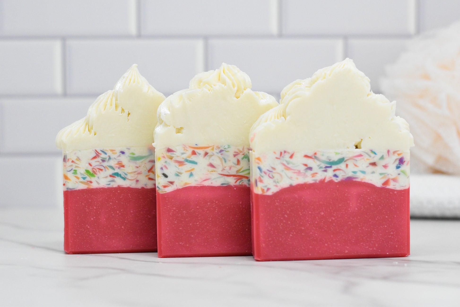 Displayed on a marble surface are three decorative bars of Birthday Cake Bar Soap from 570 Soap Co., each designed with a bright pink base and a confetti-like middle layer. The creamy white top resembles frosting and emits a delightful buttercream icing scent, set against the backdrop of white tiled walls.