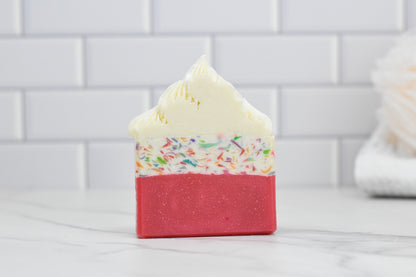 570 Soap Co.'s Birthday Cake Bar Soap showcases a red bottom layer, a middle layer adorned with colorful sprinkles, and a top that mimics whipped cream. Enhanced with Buttercream Icing Fragrance, it is artfully placed on a marble surface set against a backdrop of white tiles.