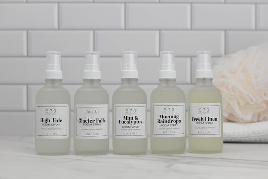 Five bottles of 570 Soap Co.'s Linen & Room Spray are arranged against a white subway tile background. The labels display the fragrances High Tide, Glacier Falls, Mint & Eucalyptus, Morning Raindrops, and Fresh Linen. Known for their natural odor-eliminating properties, these sprays provide a range of refreshing scents. To the right sits a sponge.