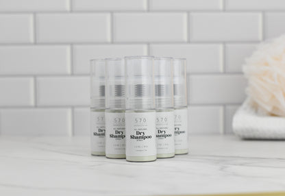 Several bottles of 570 Soap Co. Dry Shampoo, made with natural ingredients, are arranged in a neat group on a marble surface. The background showcases white subway tiles, while a soft beige towel or sponge is partially visible on the right.