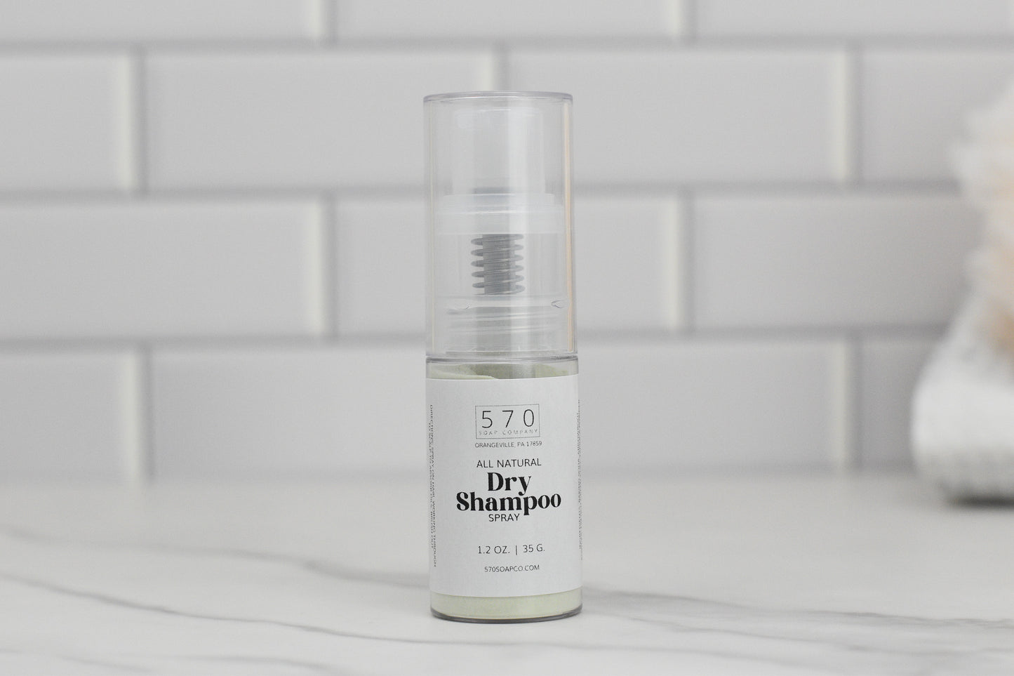 A clear bottle of all-natural dry shampoo spray from 570 Soap Co. rests on a marble surface against a white tiled backdrop. Infused with natural ingredients, the 1.2 oz (35 g) bottle promises to enhance volume while keeping your hair refreshed.