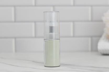 A transparent cylindrical spray bottle containing gray liquid, labeled as "Dry Shampoo" by 570 Soap Co., is set on a white marble surface. Made with natural ingredients, it enhances hair volume, while the backdrop of white subway tiles underscores a clean and minimalistic aesthetic.