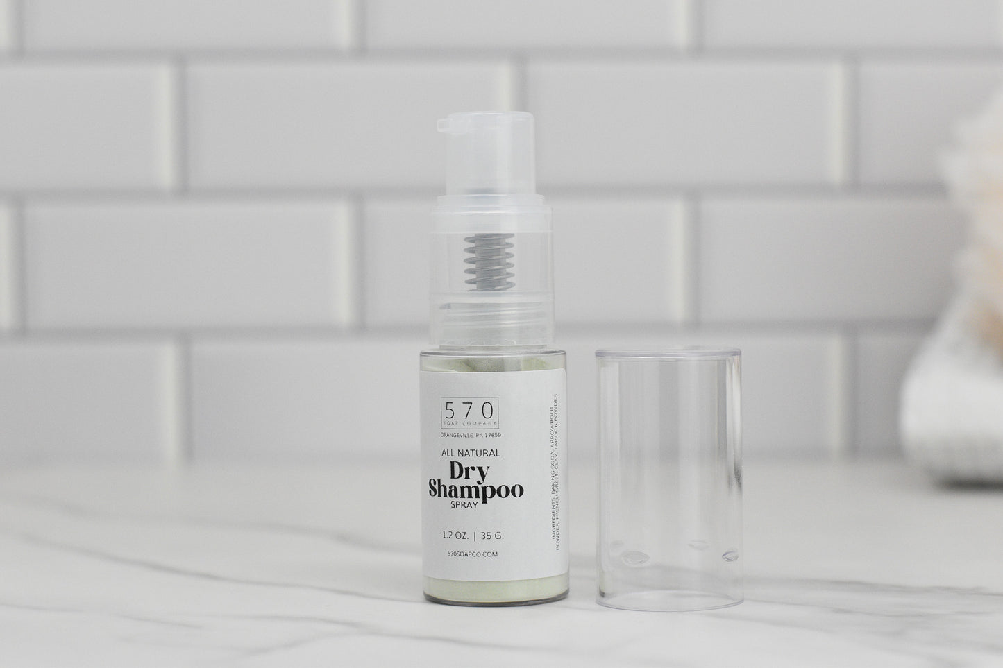 A bottle of 570 Soap Co. Dry Shampoo, renowned for its volumizing effect, is placed on a marble countertop with its transparent cap next to it against a backdrop of white tiles.