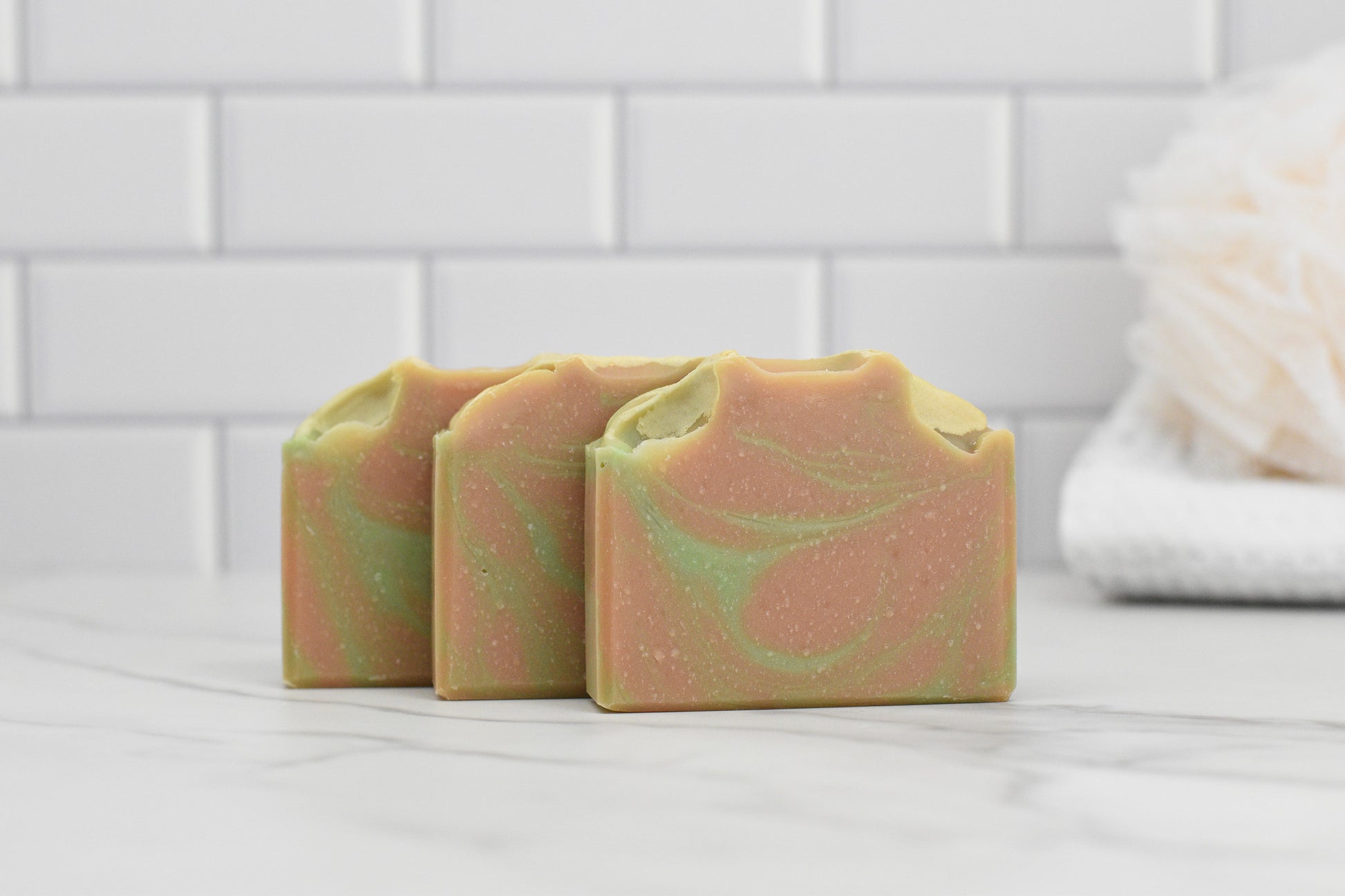 Three bars of Fresh Grapefruit & Mint Bar Soap by 570 Soap Co. are elegantly arranged on a marble surface, showcasing refreshing mint swirls and hints of grapefruit. The background features white subway tiles and a pristine white bath sponge.