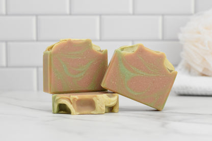 Three bars of Fresh Grapefruit & Mint Bar Soap by 570 Soap Co., featuring green and pink swirls and infused with mint and grapefruit notes, are displayed on a marble surface. A soft beige loofah is partially visible against a white tiled wall in the background.