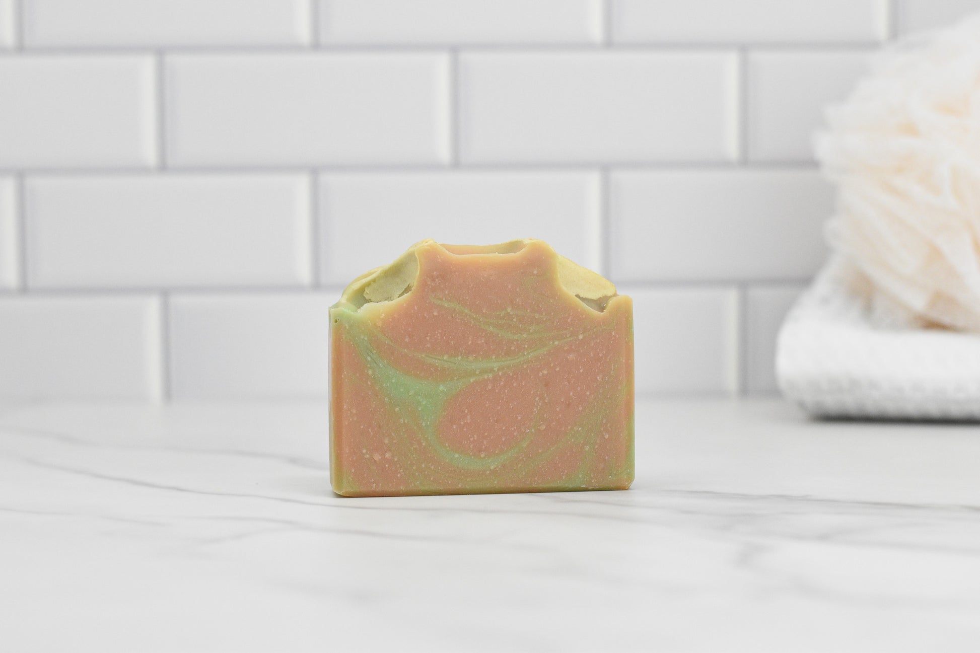 A bar of Fresh Grapefruit & Mint Soap from 570 Soap Co. showcases a swirl pattern of green, grapefruit-tinted beige, and orange as it rests on a white marble surface. The background includes white subway tiles and a cream-colored bath sponge, enhancing the freshness of your bathing experience.