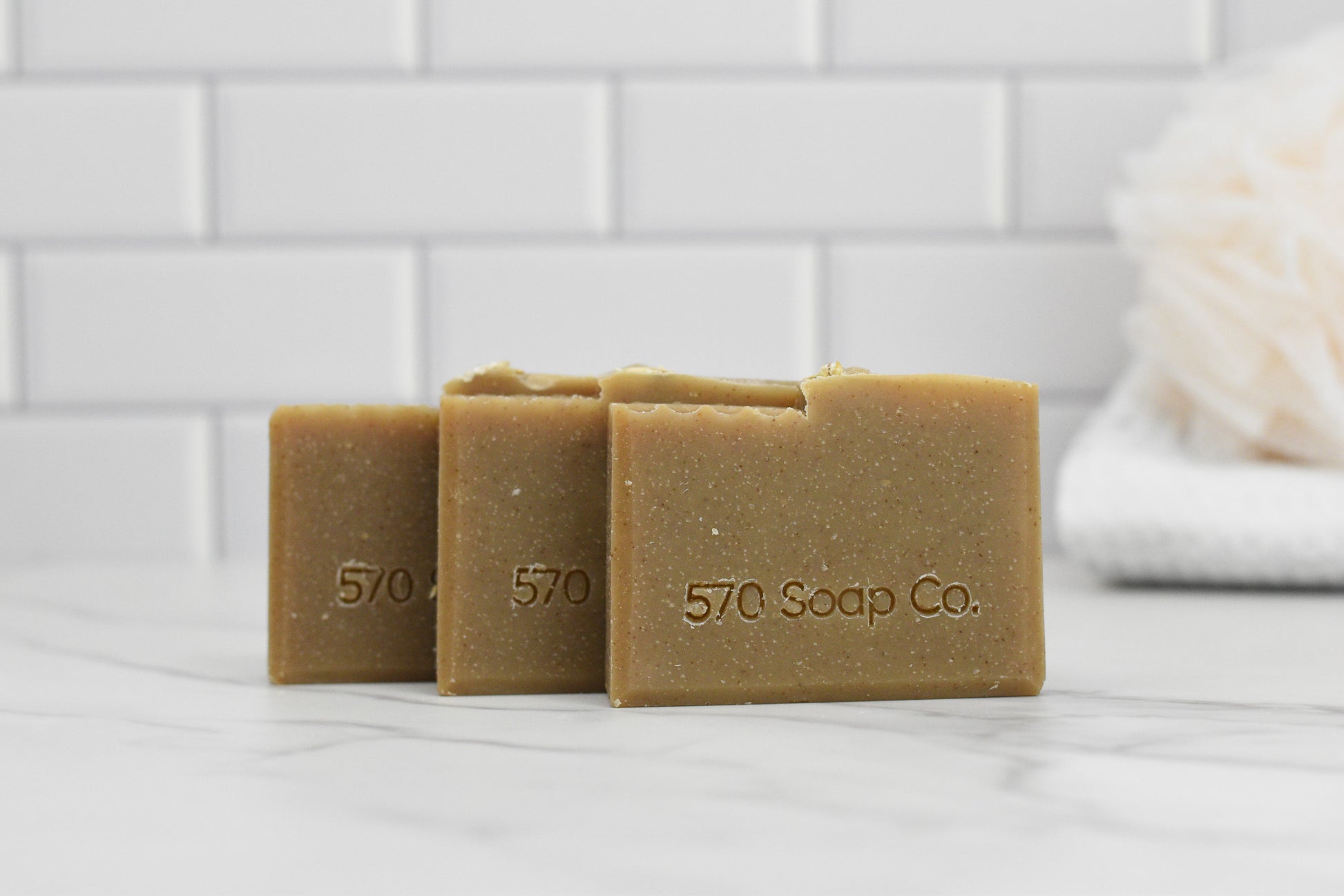 Three bars of Oatmeal Milk and Honey Bar Soap from 570 Soap Co., embossed with the brand name, are displayed on a marble surface, radiating a warm honey tone. In the background, a white loofah rests against the pristine white tile wall.