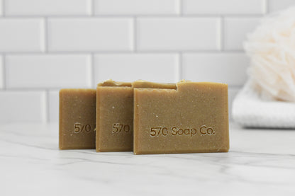 Three bars of Oatmeal Milk and Honey Bar Soap from 570 Soap Co., embossed with the brand name, are displayed on a marble surface, radiating a warm honey tone. In the background, a white loofah rests against the pristine white tile wall.