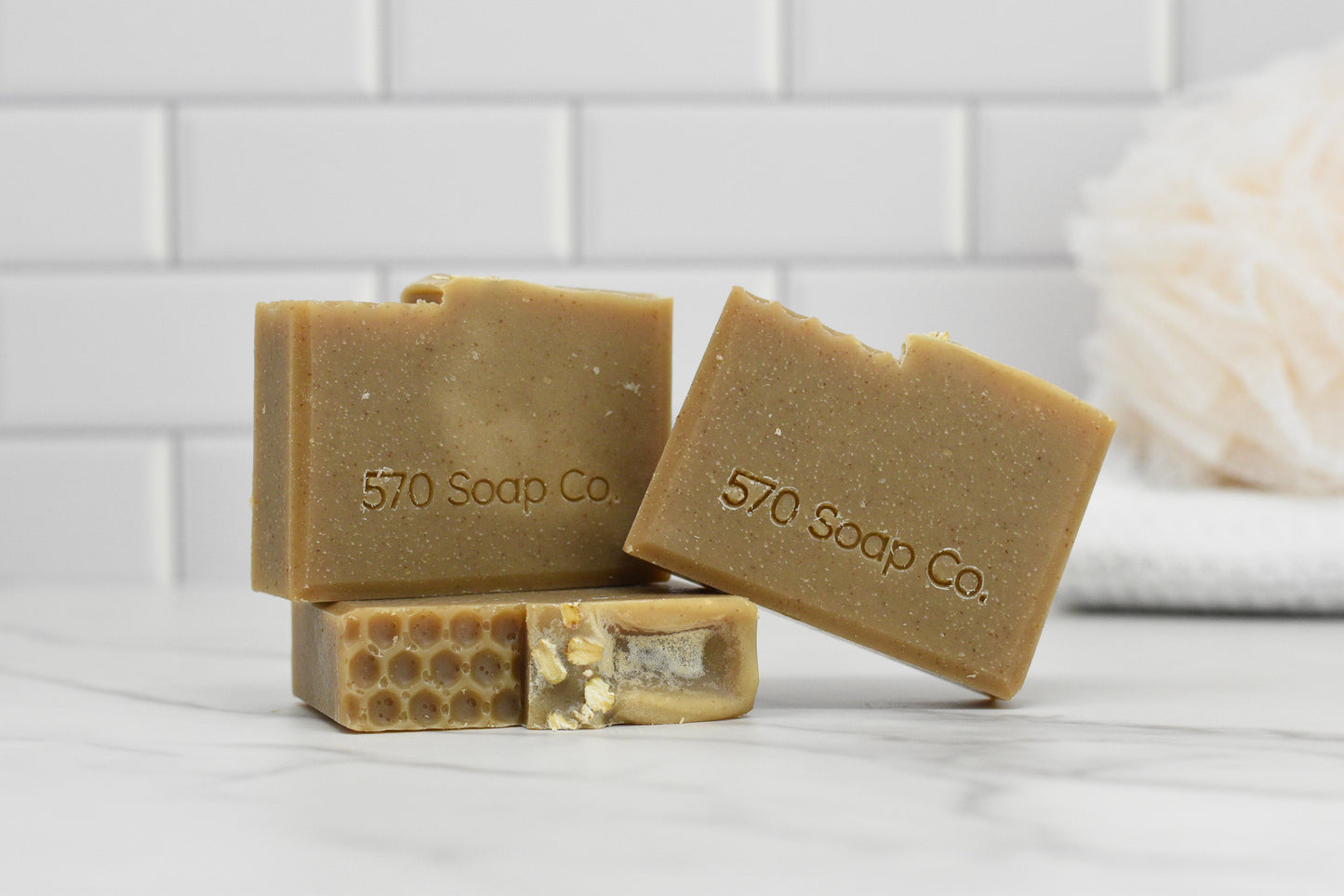 Three bars of the Oatmeal Milk and Honey Bar Soap from 570 Soap Co. are stacked on a marble surface, with one bar showcasing a honeycomb pattern indicative of its calming mix of milk and honey. In the blurred background, there's a white loofah set against stylish white subway tiles.