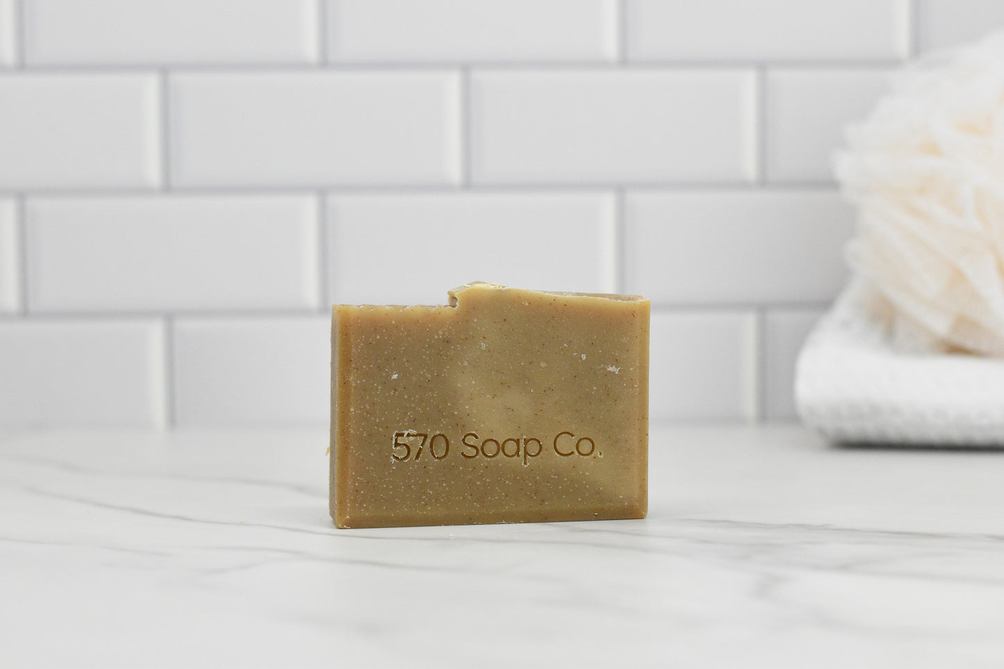 The Oatmeal Milk and Honey Bar Soap from 570 Soap Co. is elegantly displayed on a marble surface, with white subway tiles and a gentle loofah in the background, creating a clean and minimalist aesthetic.