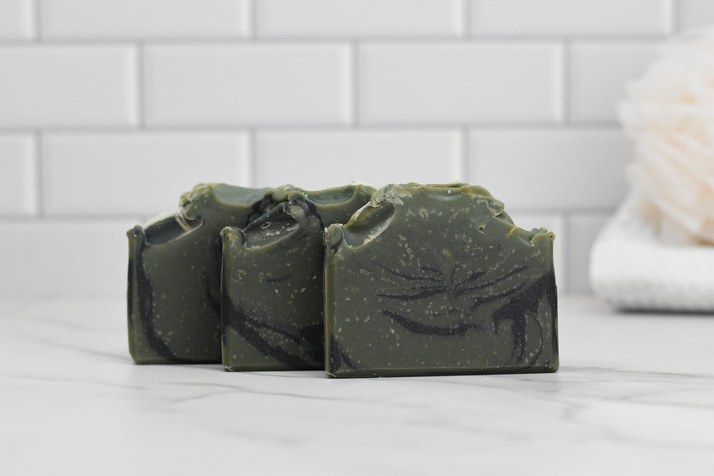 Three bars of 570 Soap Co.'s Glacier Woods Bar Soap, featuring dark green swirled patterns, are lined up on a white marble surface. In the background, there are white subway tiles, and a beige loofah is visible on the right side, adding to this elegant bathroom scene.