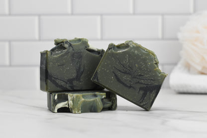 Three Glacier Woods Bar Soaps, handmade by 570 Soap Co., featuring marbled patterns, are stacked on a white marble surface. This luxurious soap captivates the scene, drawing attention against the backdrop of a white tiled wall.