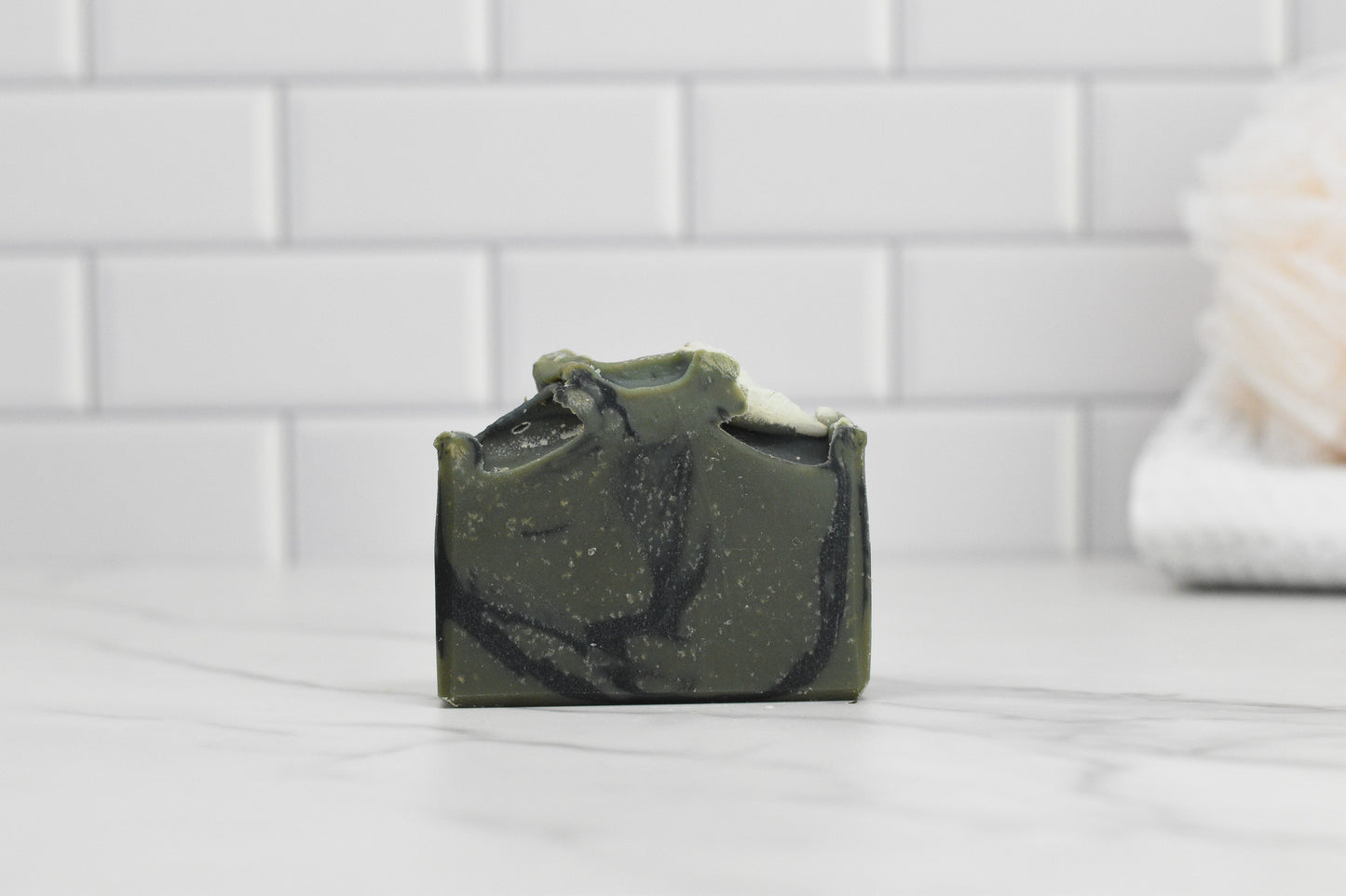 A luxurious Glacier Woods Bar Soap by 570 Soap Co., showcasing a marbled swirl of dark green and black, rests on a white marble surface. The background highlights white subway tiles and a blurred, light-colored bath sponge, creating an elegant ambiance.