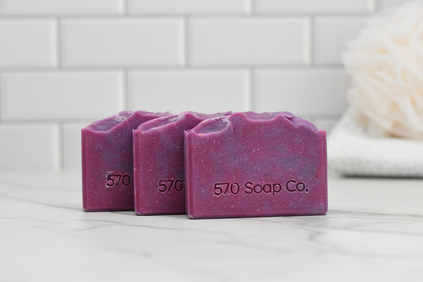 Three bars of Luscious Lilac Bar Soap: Floral by 570 Soap Co. are showcased on a marble surface. The background includes white subway tiles and a beige bath sponge, suggesting a nourishing blend with a hint of floral scent.