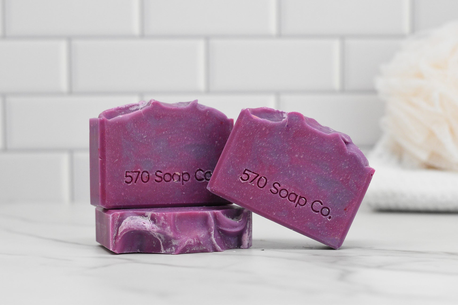 The Luscious Lilac Bar Soap by 570 Soap Co. boasts marbled patterns in purple tones and has the brand's name embossed on each bar. Exuding a delicate floral scent, these handmade soaps are showcased on a white marble surface, with a soft towel in the background against a white tiled wall, highlighting their nourishing blend of elegance and craftsmanship.