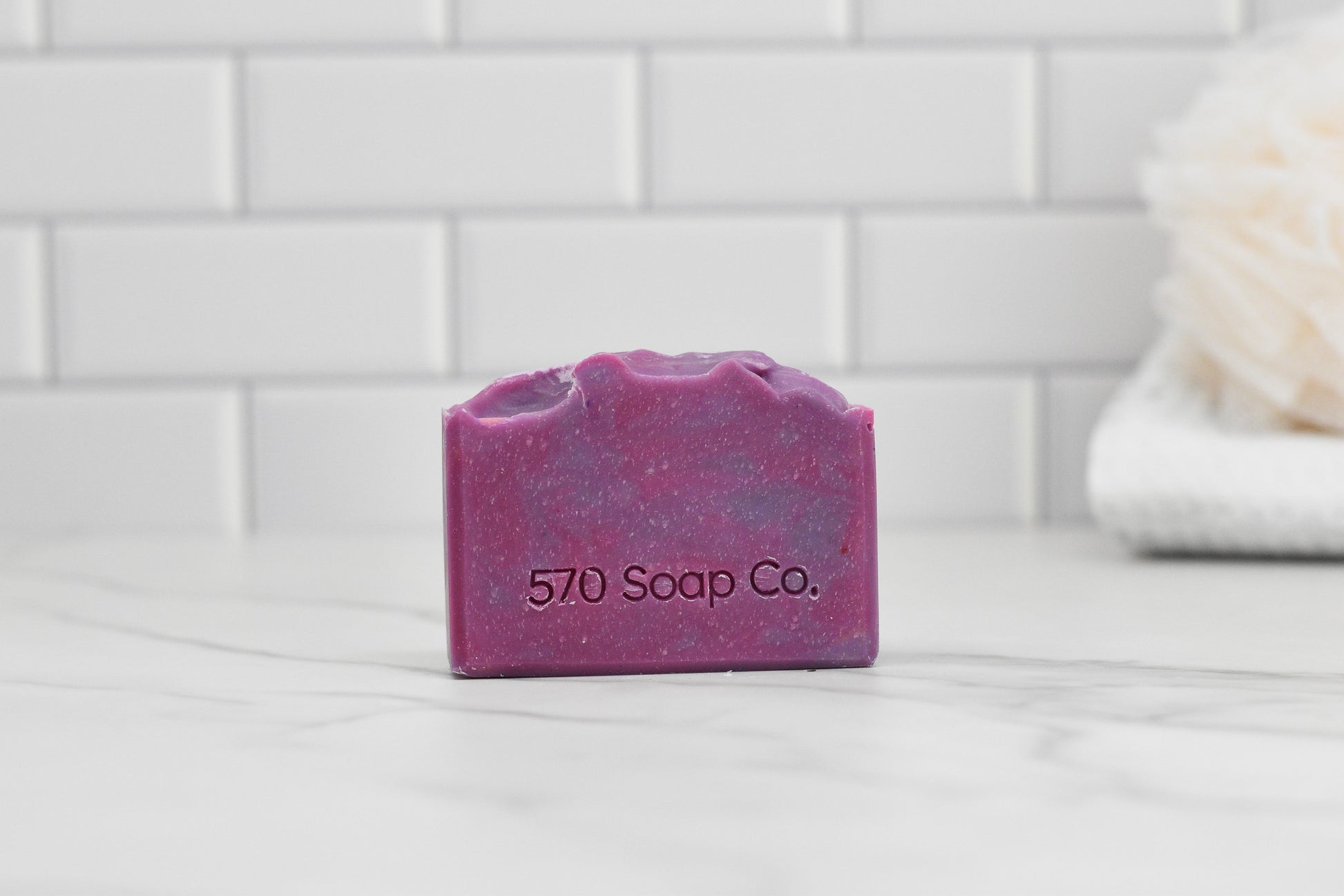 A rectangular soap bar from 570 Soap Co., embossed with the brand's name and showcasing a marbled lilac design, is set against a white tiled backdrop. Partially visible on the right is a luxurious white bath sponge, hinting at the nourishing blend of the Luscious Lilac Bar Soap: Floral.