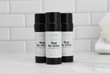 Three black cylindrical Bug-Be-Gone Lotion Sticks by 570 Soap Co. are arranged on a white marble surface with a tiled white background. These 2-ounce sticks function as an all-natural bug repellent, featuring natural ingredients that ensure effective protection.