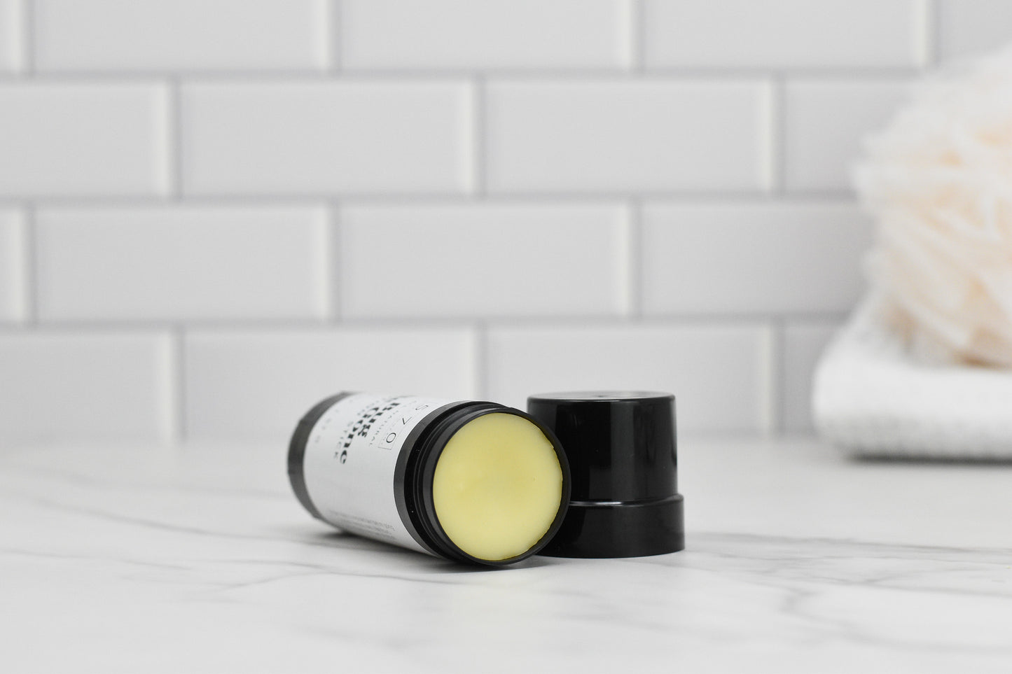 A stick of Bug-Be-Gone Lotion Stick from 570 Soap Co., featuring a black cap and made with natural ingredients, lies open on a marble surface. The background showcases white subway tiles and a blurred loofah resting on a folded towel.