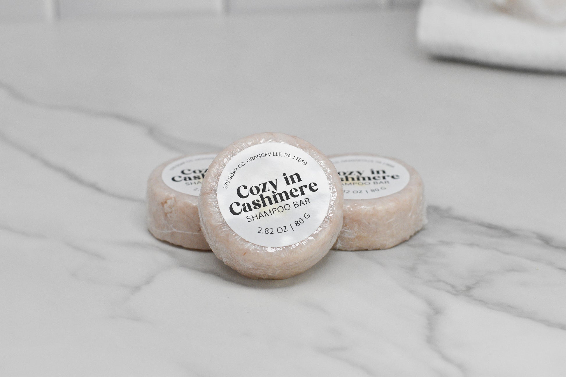 Three Cozy in Cashmere Shampoo Bars from 570 Soap Co. are elegantly displayed on a marble surface. Enclosed in light-colored packaging, these eco-friendly bars provide luxurious hair care. The softly blurred white-tiled wall in the background enhances their minimalist allure.