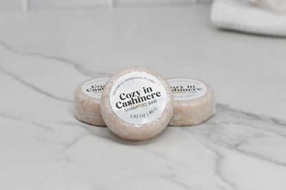 Three Cozy in Cashmere Shampoo Bars from 570 Soap Co. are elegantly displayed on a marble surface. Enclosed in light-colored packaging, these eco-friendly bars provide luxurious hair care. The softly blurred white-tiled wall in the background enhances their minimalist allure.