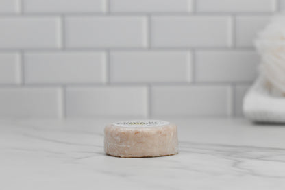 The Cozy in Cashmere Shampoo Bar by 570 Soap Co. sits elegantly on the marble countertop, framed by a white subway tile backsplash. Infused with nourishing ingredients, this eco-friendly hair care essential promises a lush experience. A blurred loofah peeks into view on the right.