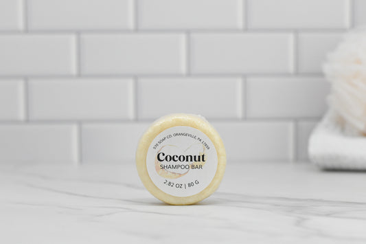 A round Coconut Shampoo Bar from 570 Soap Co., infused with a tropical coconut fragrance, is displayed on a marble surface. The backdrop highlights white subway tiles and, to the right, a softly blurred object resembling a bath sponge. The shampoo bar's label indicates it weighs 2.82 oz / 80 g, making it perfect for eco-friendly hair care.