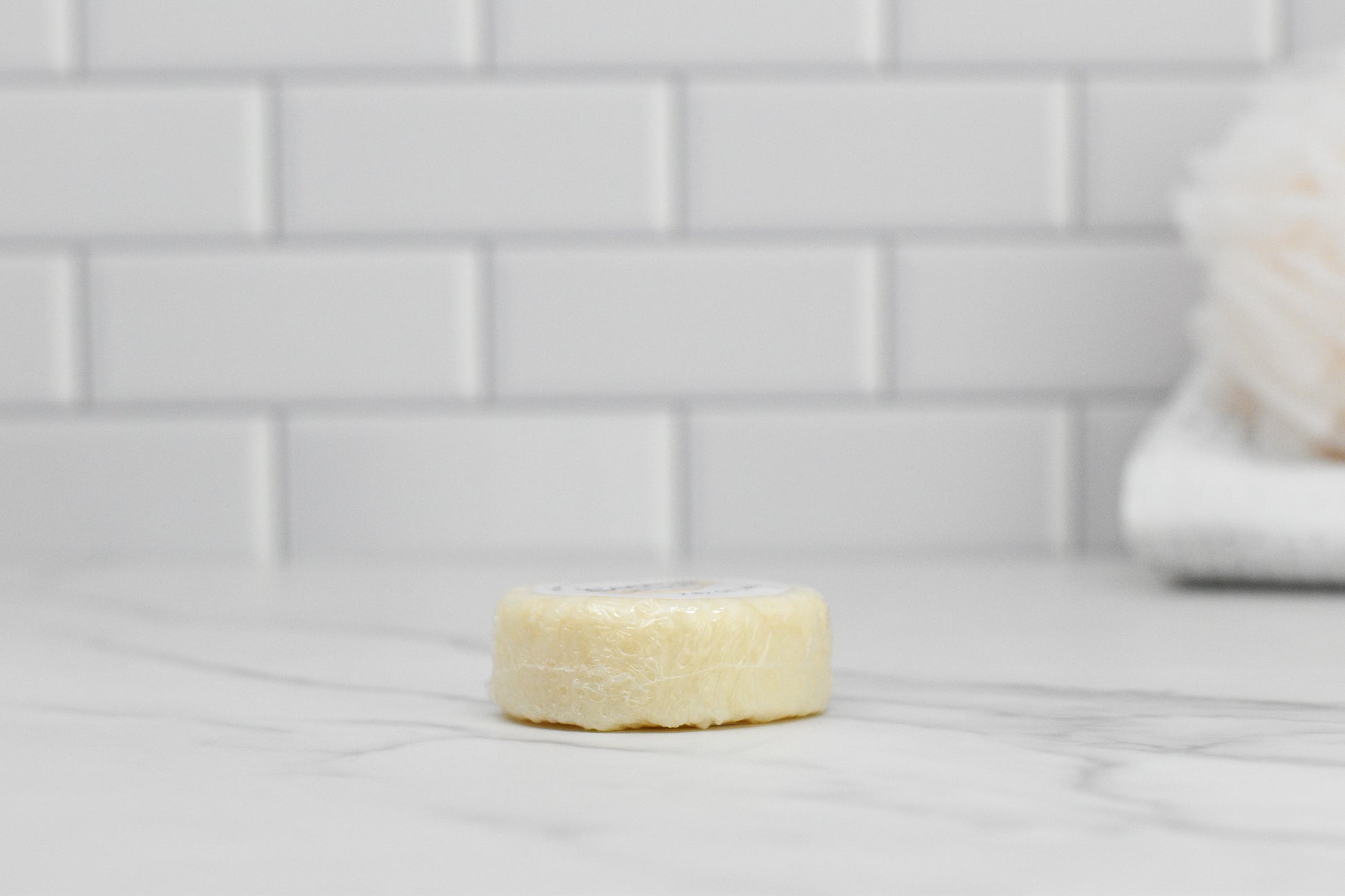 A small, round Coconut Shampoo Bar from 570 Soap Co. rests on a marble countertop. The setting includes white, subway-style tiled walls and a blurred, fluffy bath sponge in the background, conveying an eco-friendly hair care sanctuary.