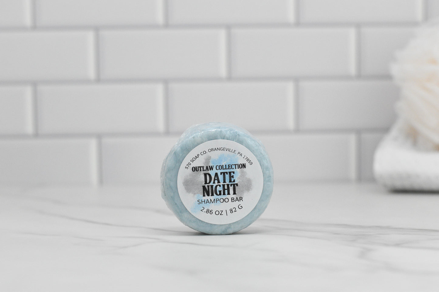 A luxurious hair care gem, the Date Night Shampoo Bar from 570 Soap Co. graces a white countertop against a tiled backsplash. This eco-friendly shampoo bar is round, light blue, and weighs 2.86 ounces (92 grams), making it perfect for an indulgent cleansing experience.