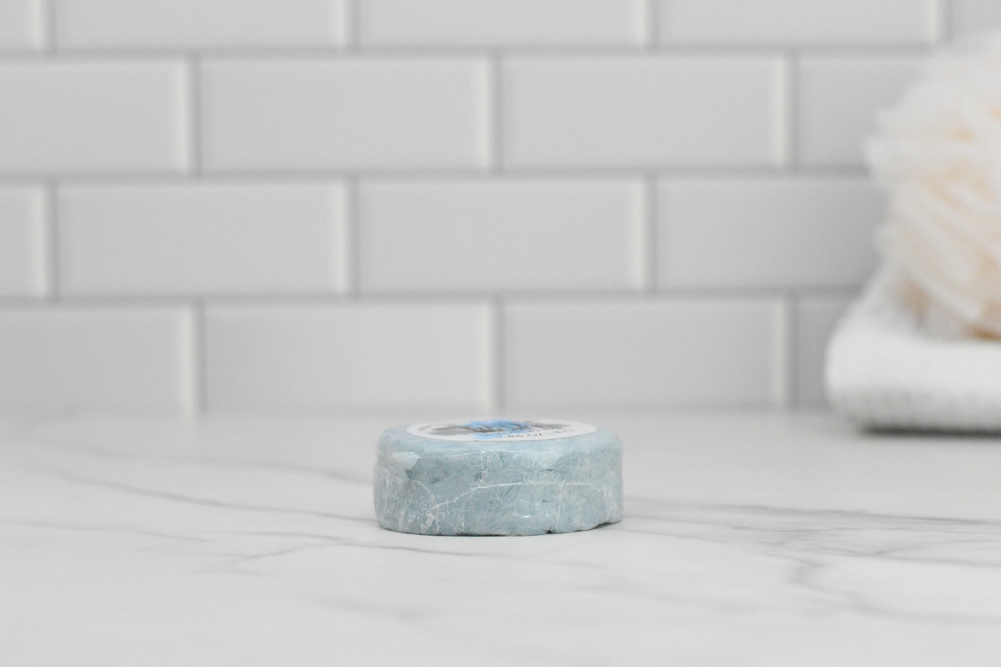 The Date Night Shampoo Bar from 570 Soap Co., a round blue eco-friendly product, is showcased on a marble countertop with a white subway tile backsplash. In the background, a fluffy bath accessory is slightly blurred, enhancing the elegant ambiance ideal for luxurious hair care.