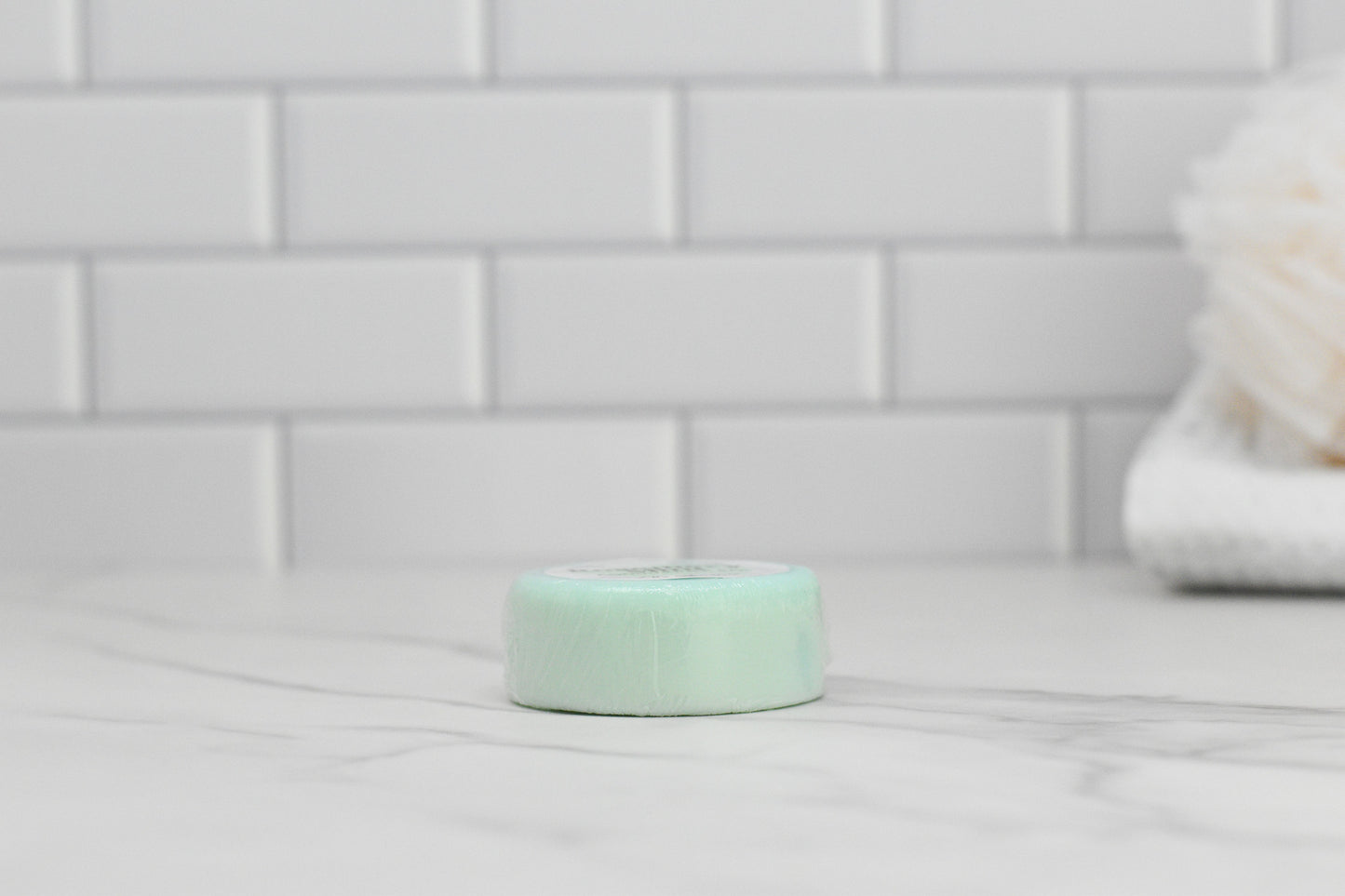 A light green bar of the Extra Nourishing Conditioner from 570 Soap Co., infused with Rosemary & Peppermint Essential Oil, sits on a marble countertop against a backdrop of white subway tiles, with a blurred white bath sponge in the background, suggesting an eco-friendly hair care routine.