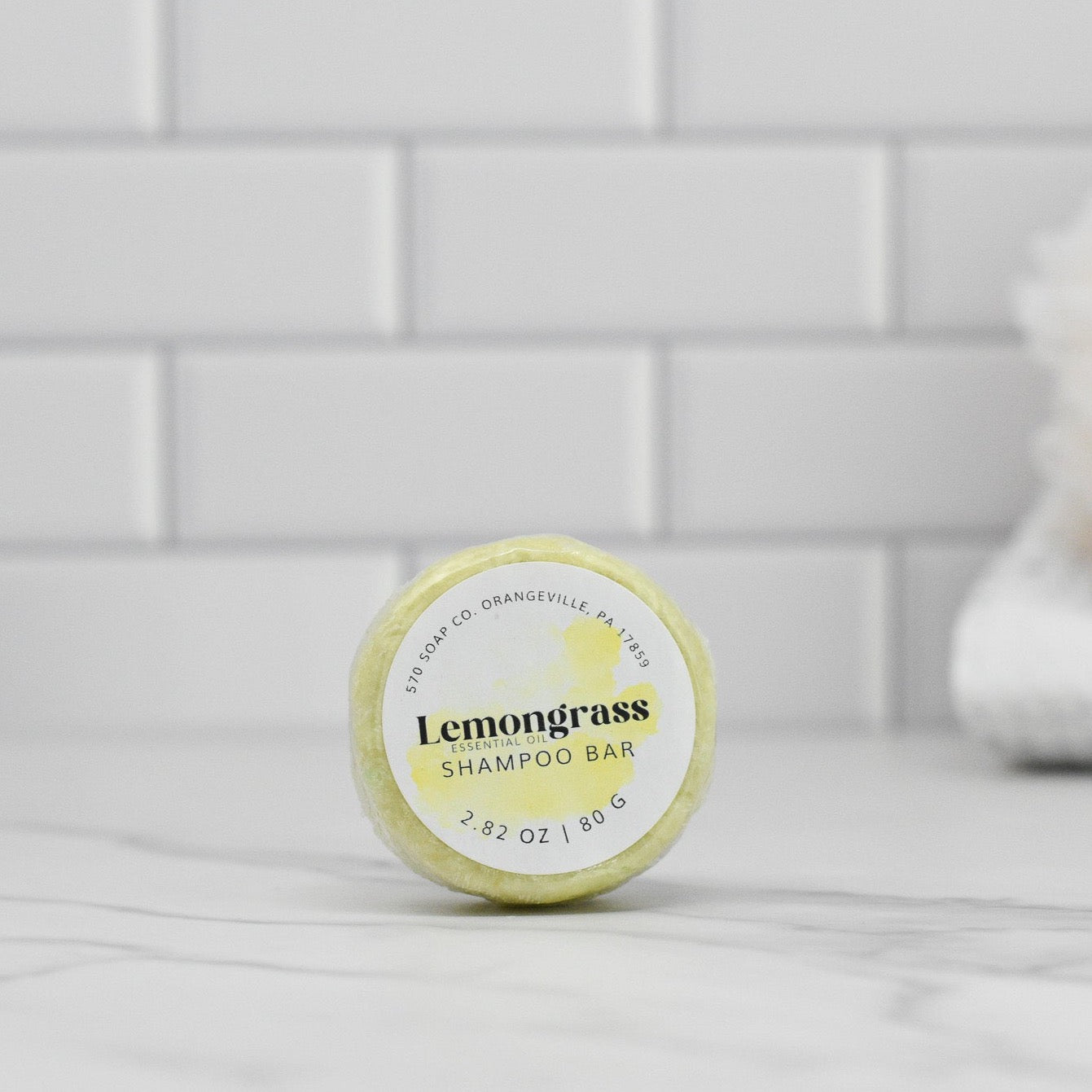 The Lemongrass Essential Oil Shampoo Bar by 570 Soap Co. sits on a marble surface against white subway tiles. The circular bar, in clear packaging with a labeled yellow accent, is suitable for all hair types. A soft, blurred white towel is visible in the background.