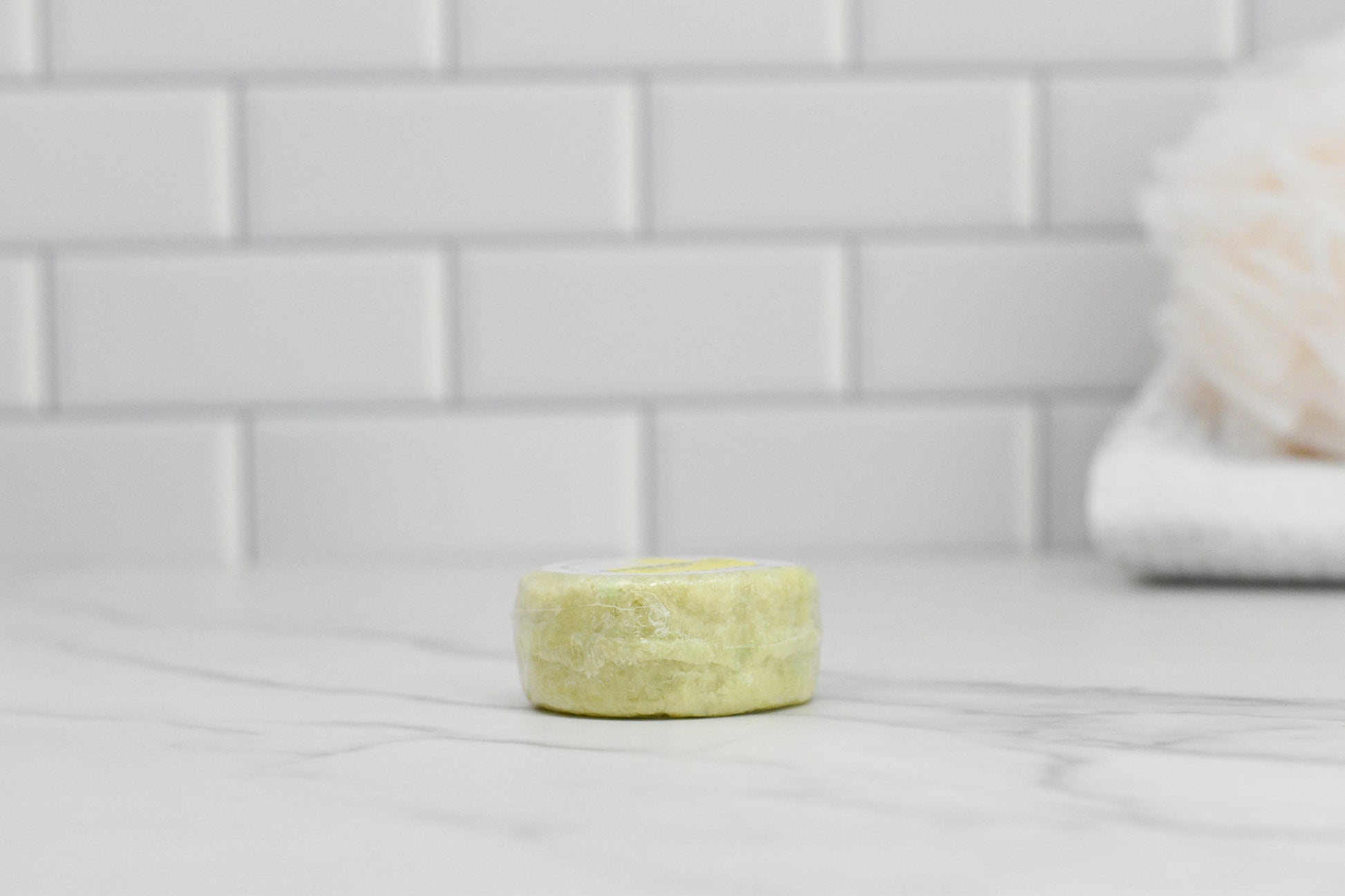 On a marble countertop, a round, pale green 570 Soap Co. Lemongrass Essential Oil Shampoo Bar sits enriched with lemongrass. A white subway tile wall appears in the background with a blurred textured object to the right. Suitable for all hair types.