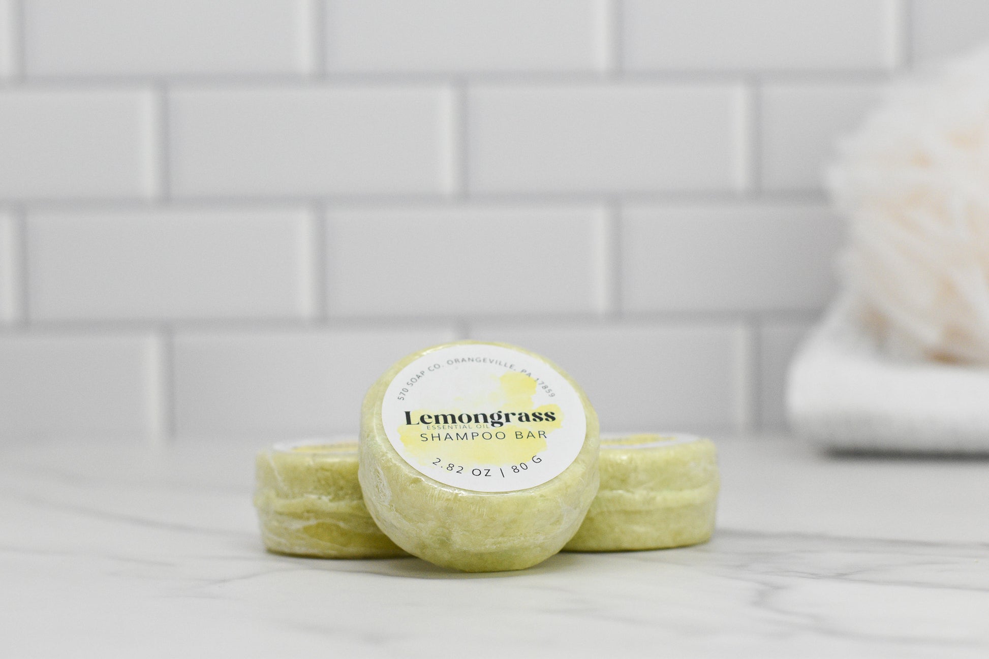 Three Lemongrass Essential Oil Shampoo Bars from 570 Soap Co. are stacked on a marble countertop in front of a white tile backsplash, with a soft towel visible in the background. Suitable for all hair types, these bars are infused with lemongrass essential oil.