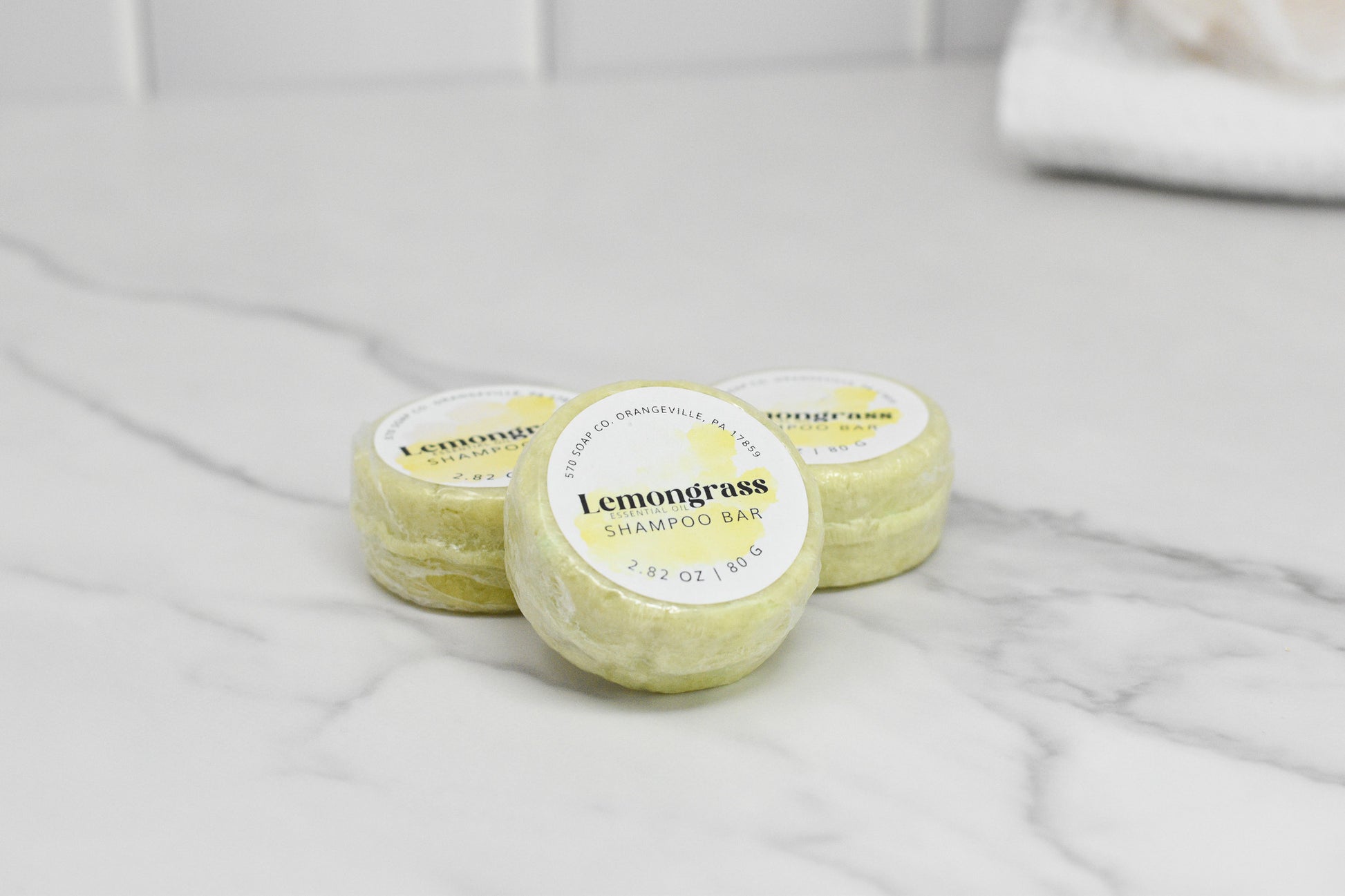 Three round 570 Soap Co. Lemongrass Essential Oil Shampoo Bars sit on a white marble surface, each labeled for scent and weight, suitable for all hair types. A white towel is partially visible in the background.