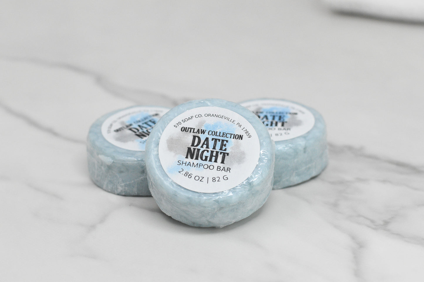 Three eco-friendly Date Night Shampoo Bars from 570 Soap Co. are elegantly displayed on a light gray marble surface. Each round blue bar, weighing 2.86 oz (82 g), offers luxurious hair care.