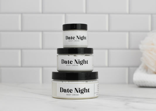 Three jars of Date Night Body Butter by 570 Soap Co., enriched with shea butter, are stacked against a white tiled background. A loofah is partially visible on the right side.