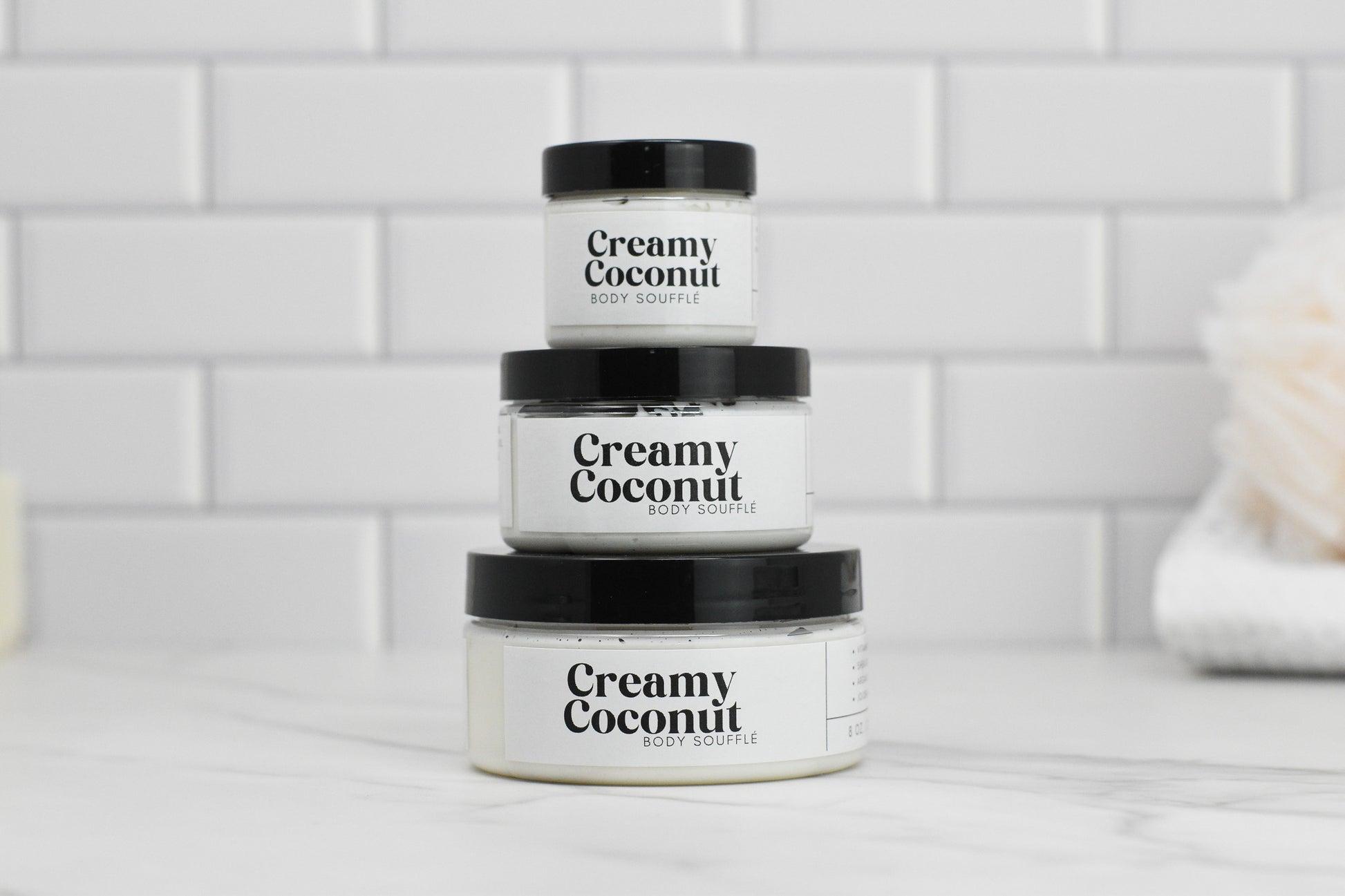 Three jars of 570 Soap Co.'s Creamy Coconut Body Butter, known for its intense hydration, are stacked against a white subway tile background. The jars come in different sizes, with the largest at the bottom and the smallest on top. A softly blurred object in the background evokes a sense of tropical paradise.