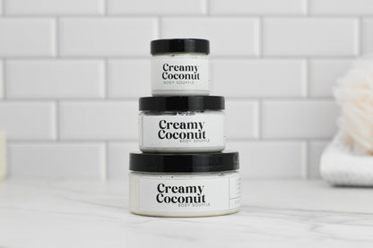 Three jars of 570 Soap Co.'s Creamy Coconut Body Butter, known for its intense hydration, are stacked against a white subway tile background. The jars come in different sizes, with the largest at the bottom and the smallest on top. A softly blurred object in the background evokes a sense of tropical paradise.